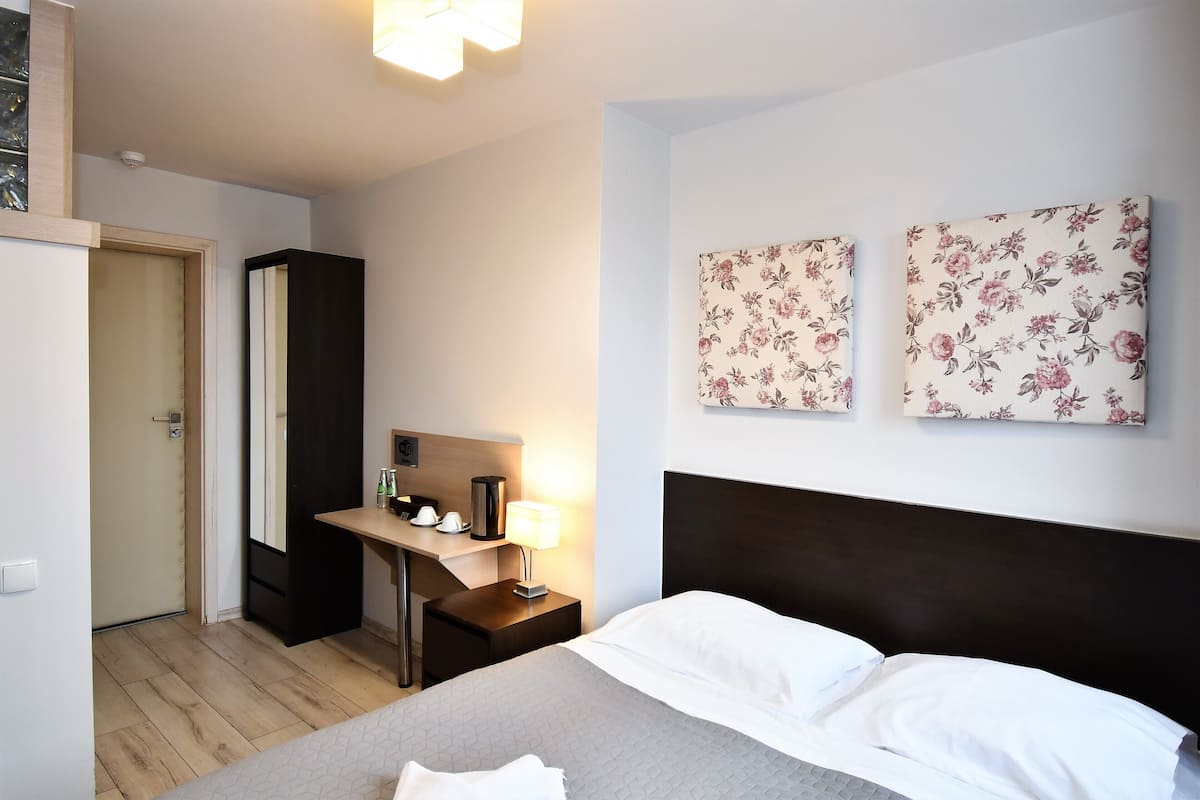 Una habitación de hotel con cama, escritorio con lámpara, armario y dos cuadros florales.