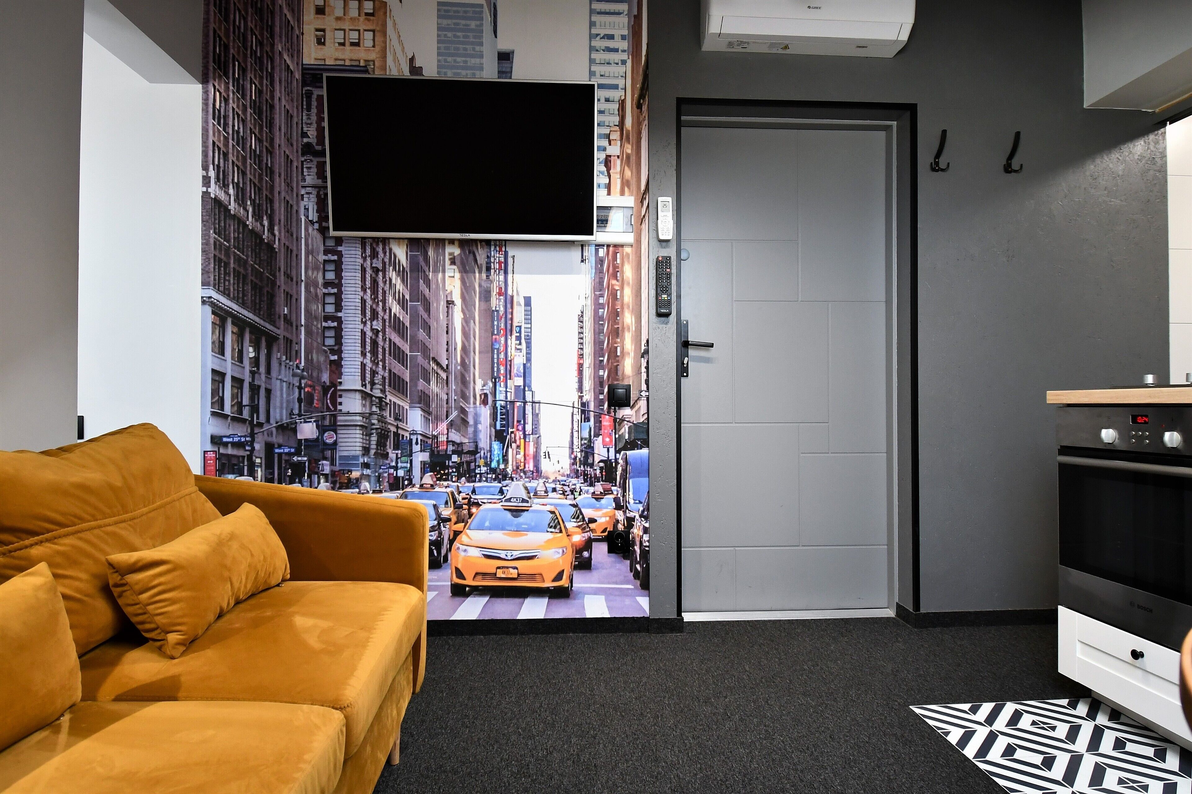 Una habitación con un amplio mural en la pared que representa una calle urbana concurrida, un sofá, una puerta y una zona de kitchenette.