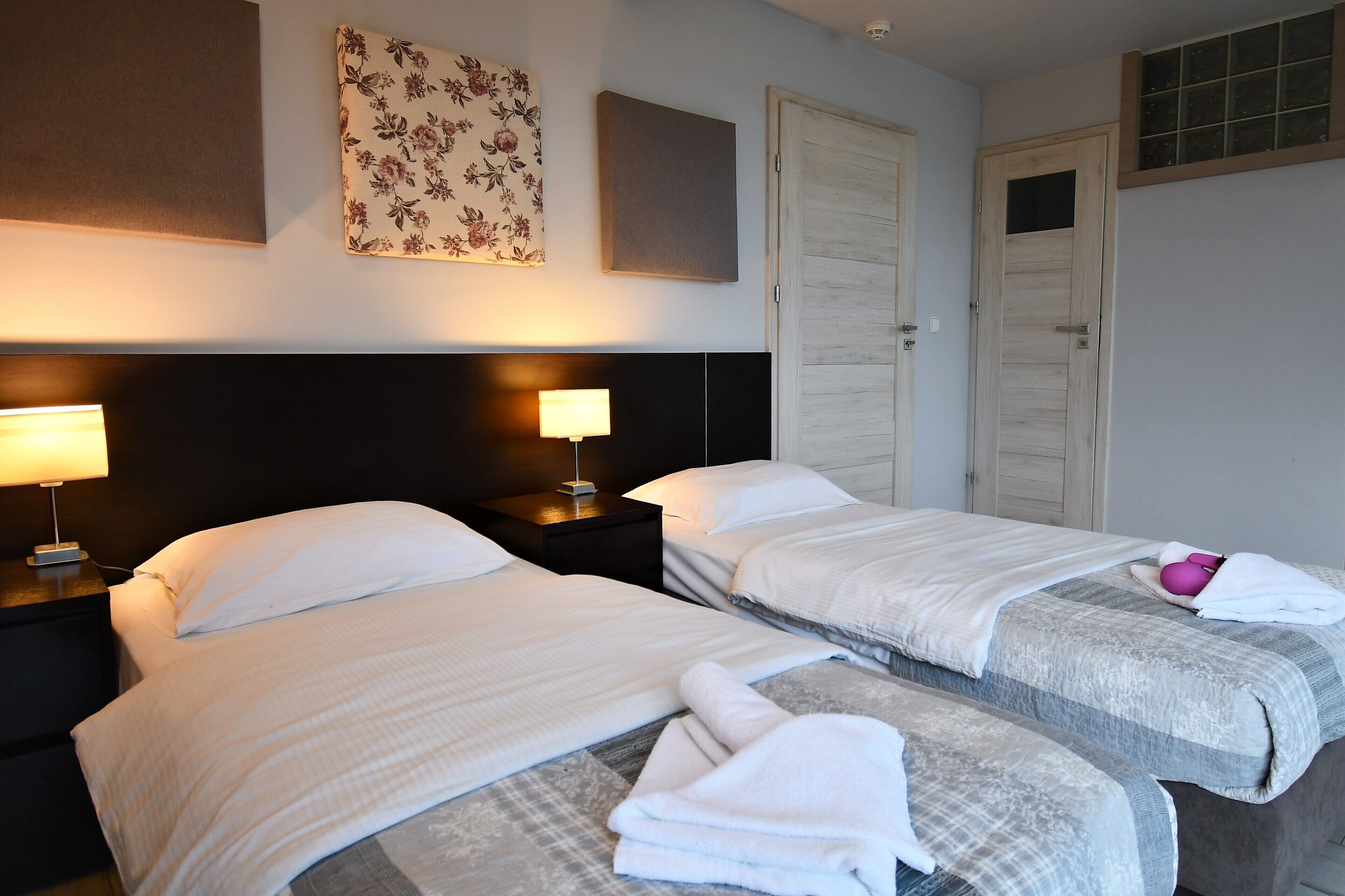 Habitación de hotel con dos camas, una mesita de noche con lámpara, una obra de arte floral en la pared y una puerta con ventanal.