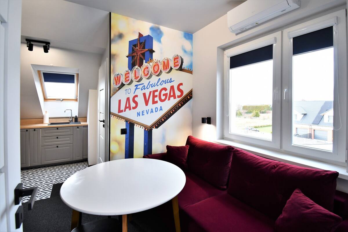 Una habitación con un cartel que dice 'Welcome to Fabulous Las Vegas Nevada', un sofá rojo, una mesa blanca, una zona de cocina y ventanas grandes.
