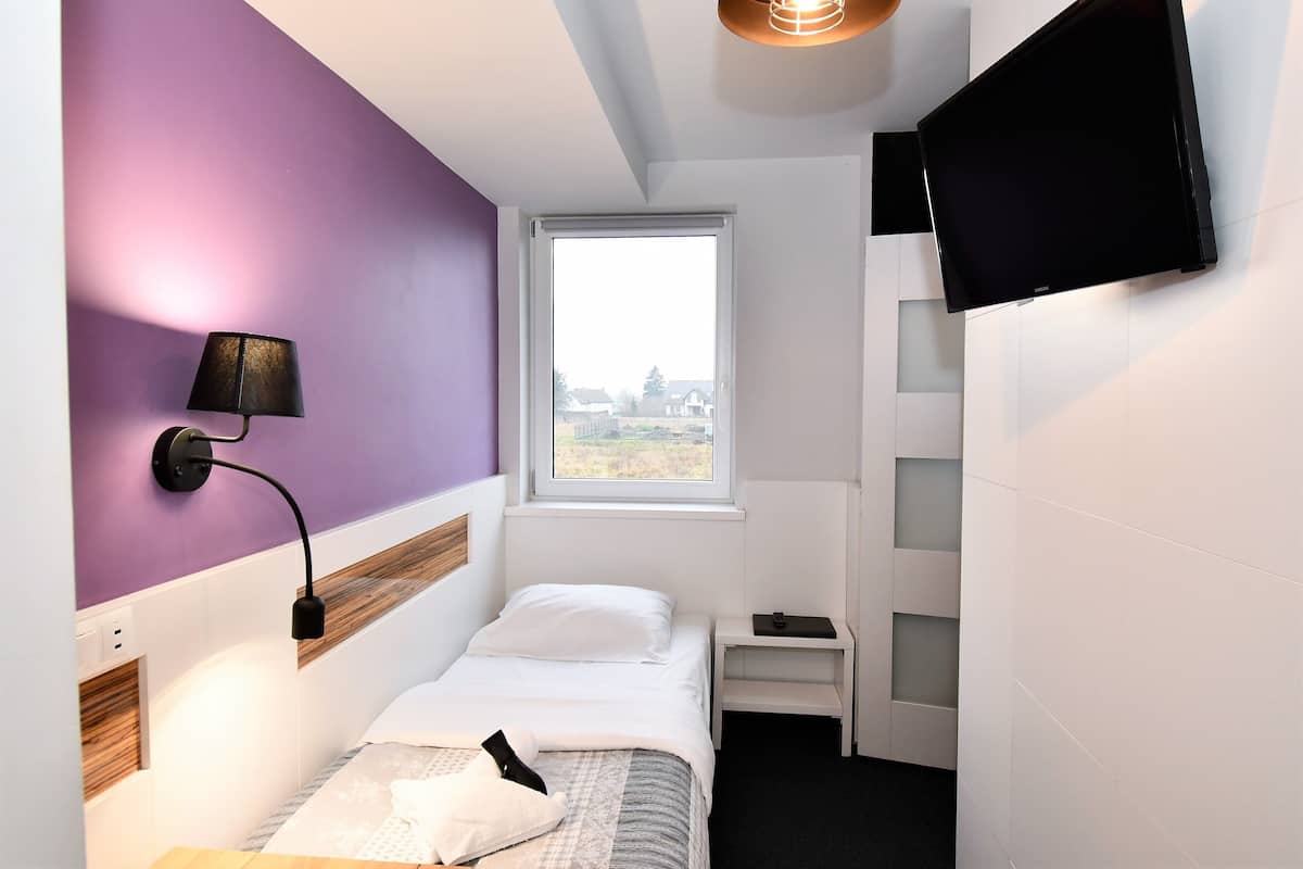Un dormitorio pequeño y moderno con cama, mesita de noche, lámpara, televisor montado en la pared y ventana con vistas.