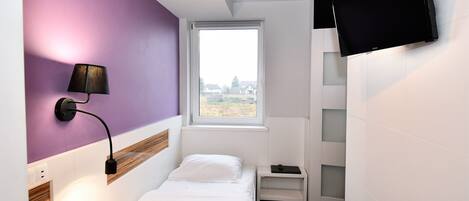 A small, modern bedroom with a bed, a bedside table, a lamp, a TV mounted on the wall, and a window with a view.