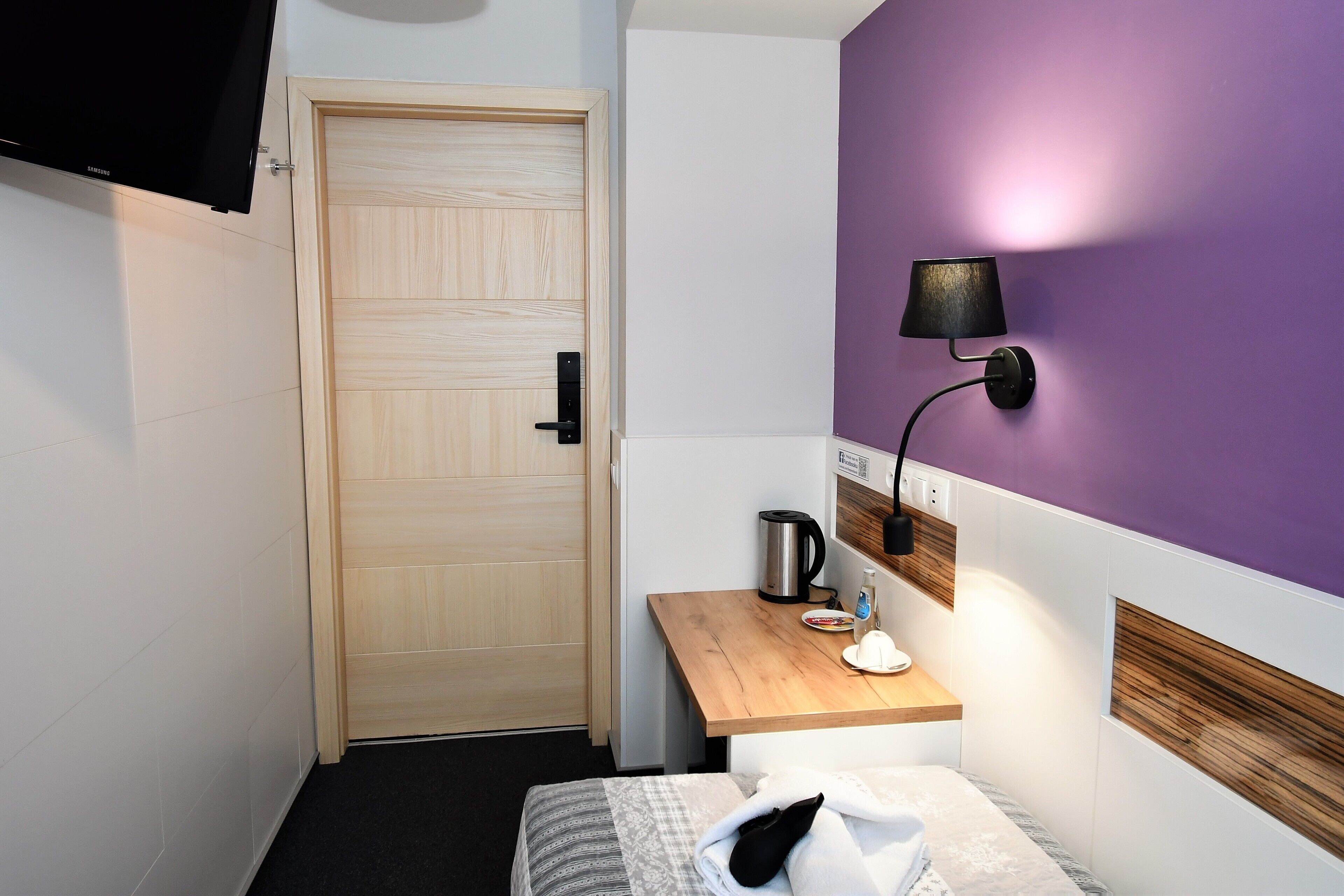 Una habitación de hotel pequeña con una puerta de madera, una mesita de noche con lámpara, una cama y una pared de acento color púrpura.
