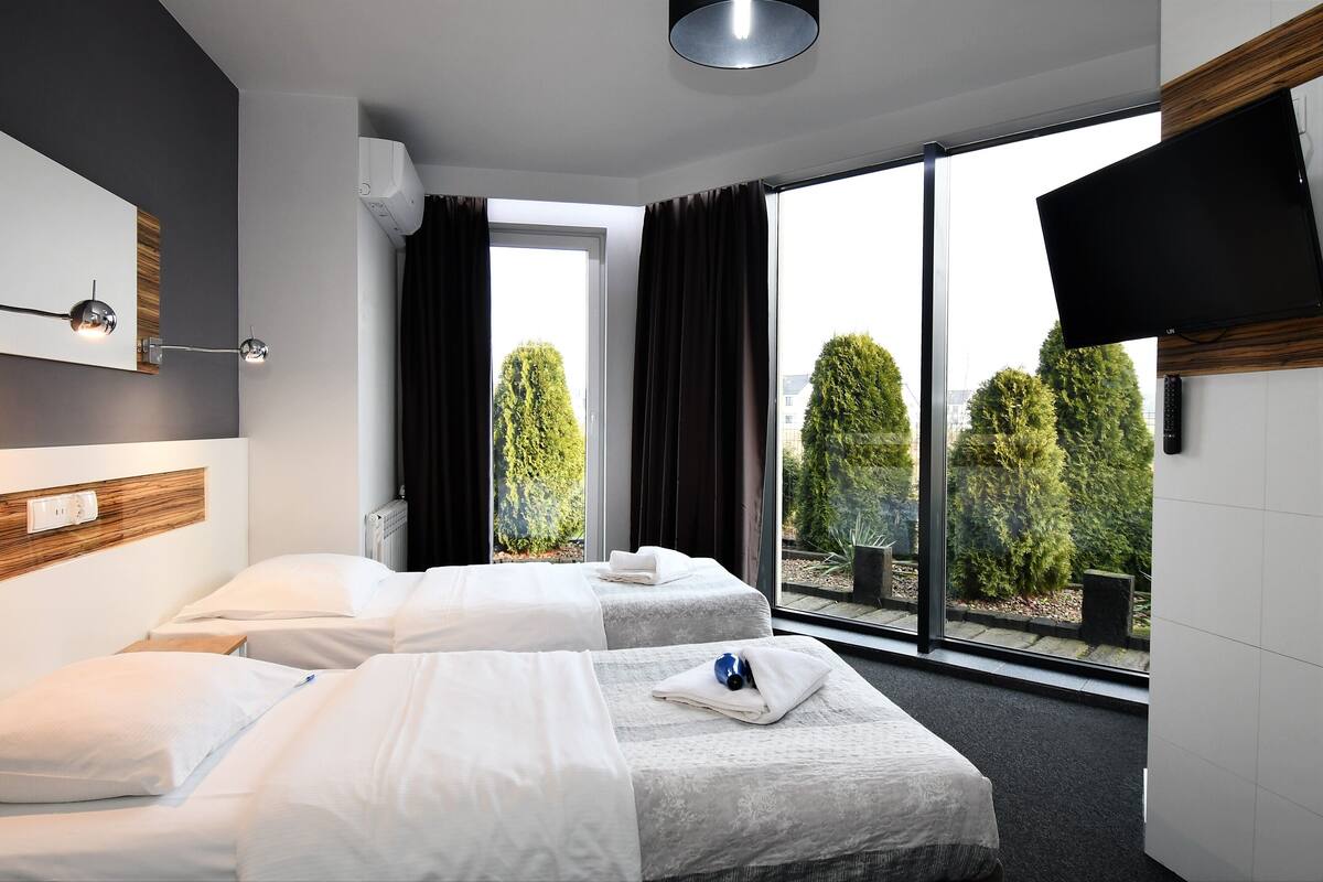 Habitación de hotel con dos camas, un ventanal con vistas a árboles, un televisor de pantalla plana montado en la pared y una lámpara en el techo.