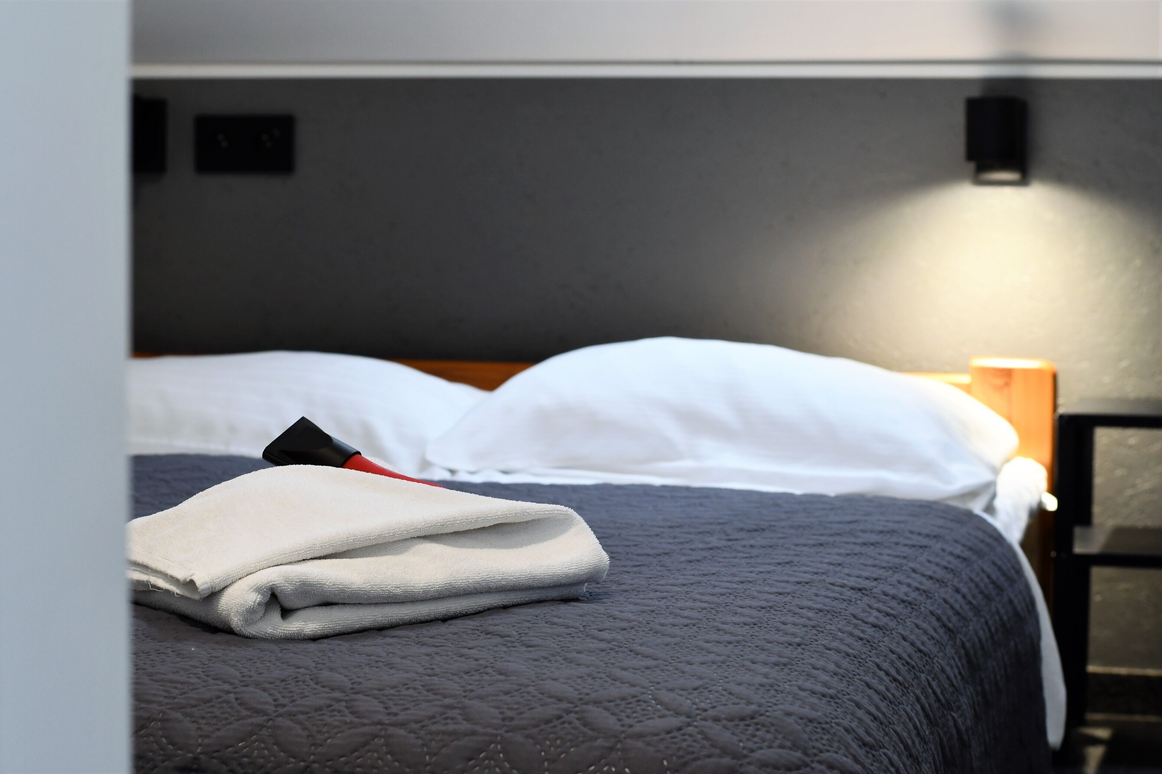 A neatly made bed with white pillows and a folded white towel, a dark quilted bedspread, and a bedside table with a lamp.