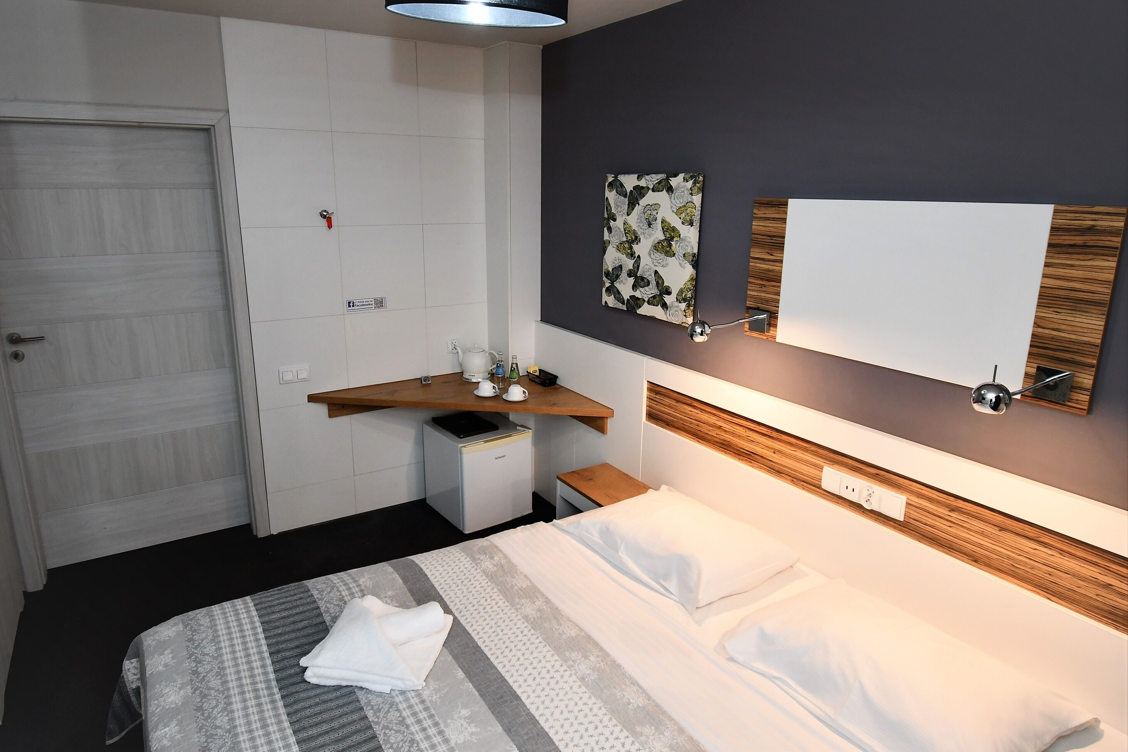 Una habitación de hotel moderna con cama, mesita de noche y un pequeño escritorio con silla. Se observa una puerta, un espejo montado en la pared y un adorno mural.