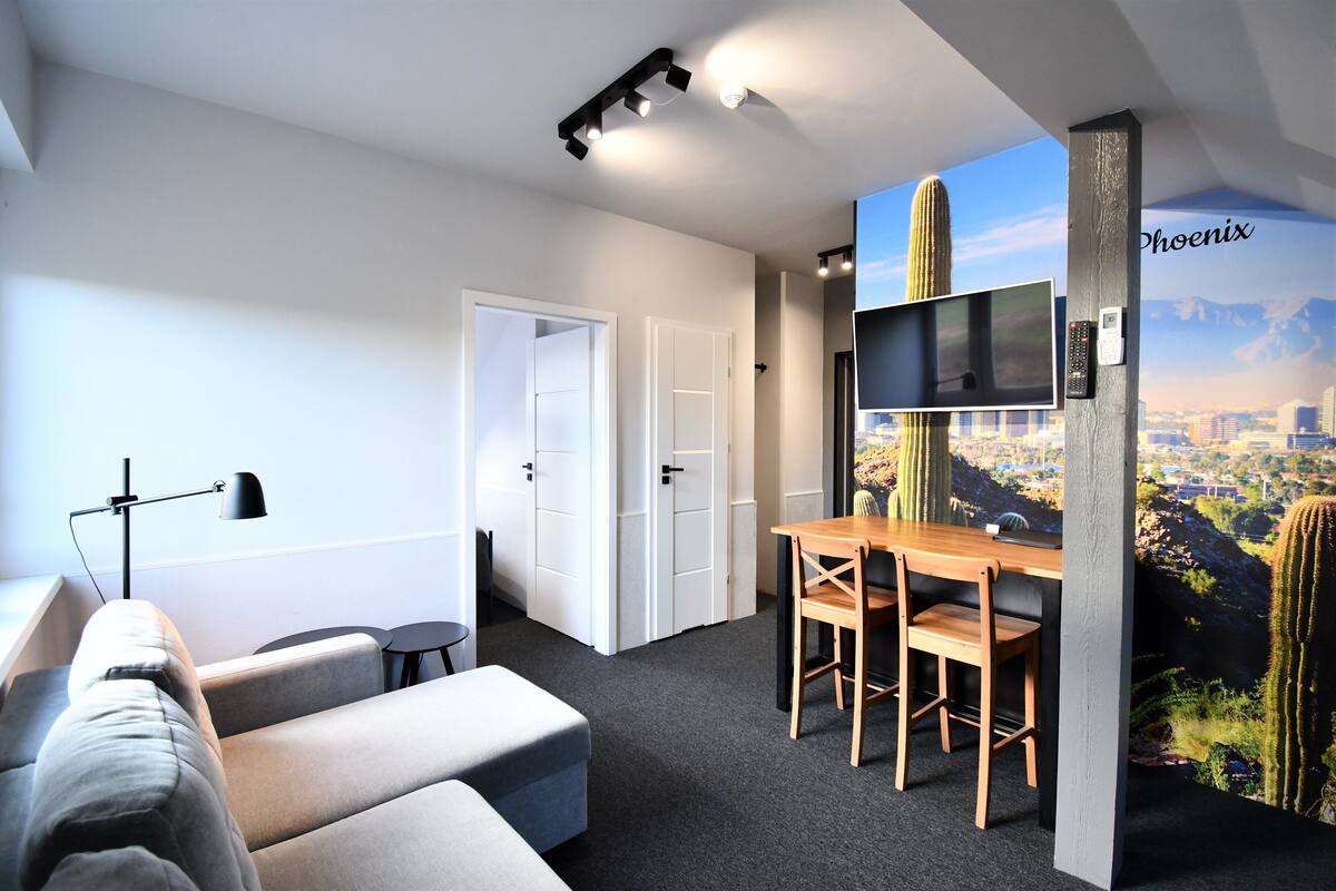 Una habitación de hotel moderna con un televisor de pantalla plana montado en la pared, una mesa de comedor de madera con sillas, un sofá, un escritorio con lámpara y una puerta que da a otra habitación.