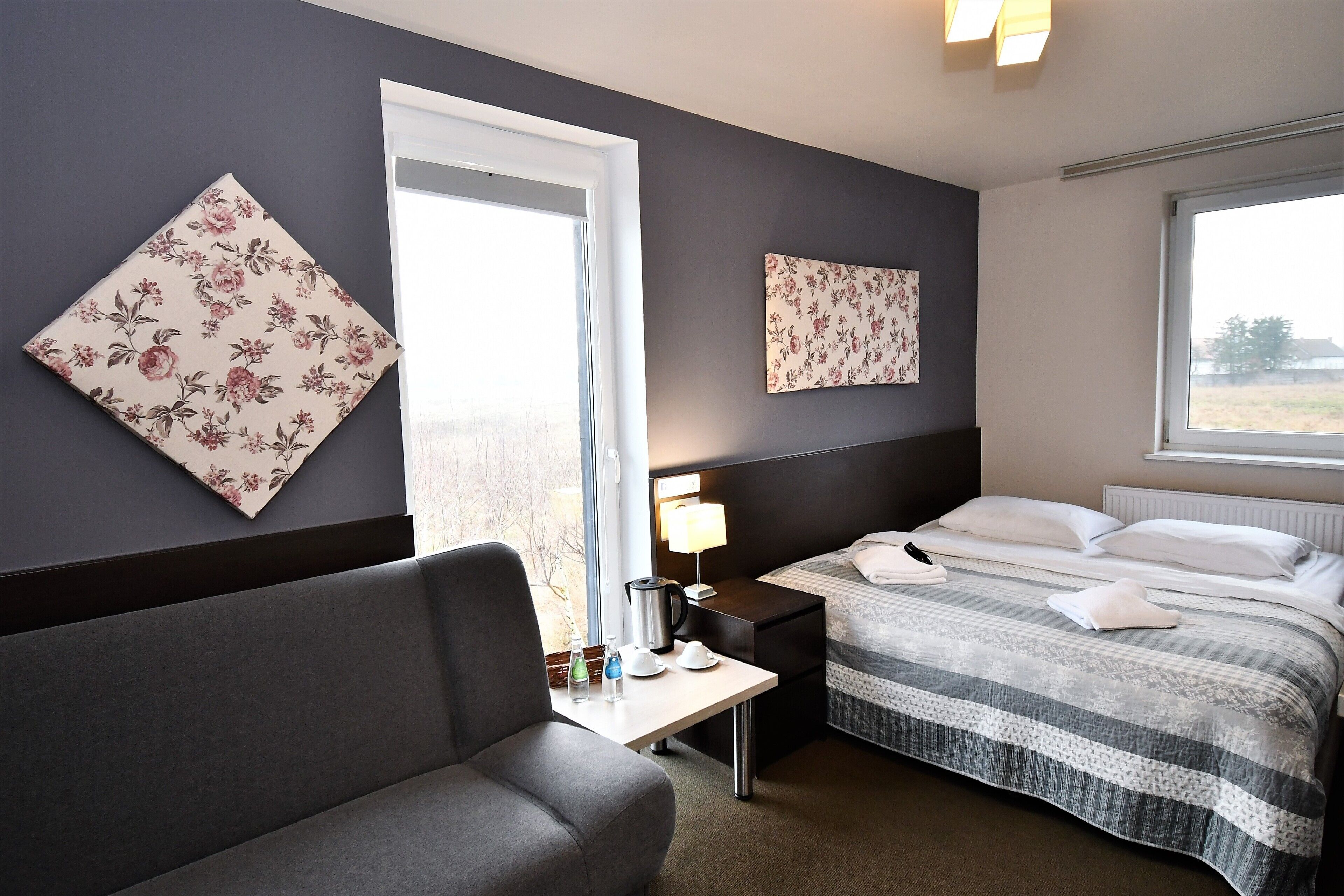 Una habitación de hotel con cama, mesita de noche, lámpara y un amplio ventanal.