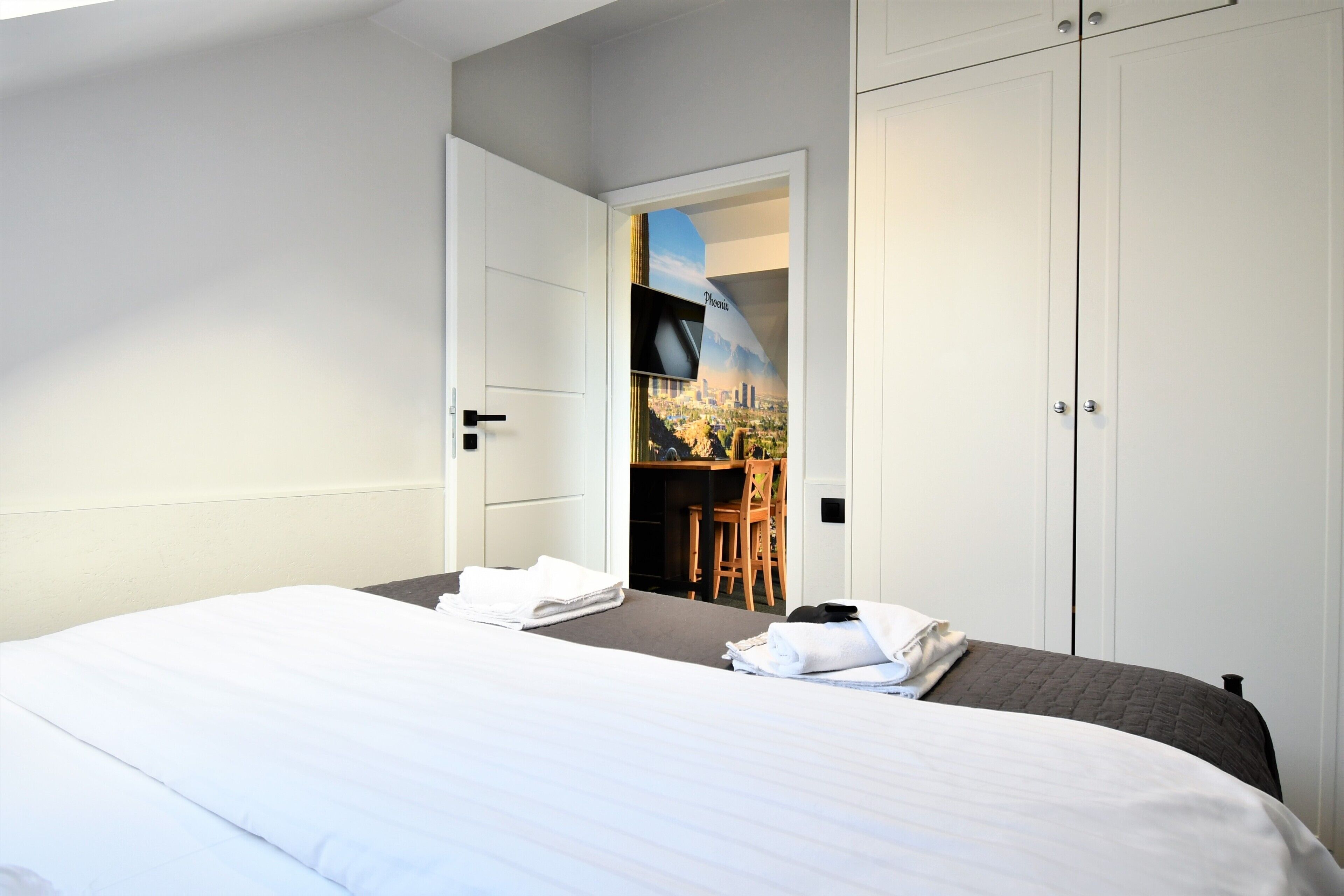 Een hotelkamer met een bed, een bureau en een stoel. Een deur leidt naar een balkon met uitzicht op de stad.