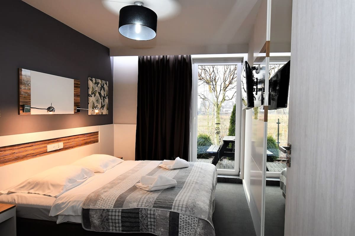 Una habitación de hotel moderna con una cama grande, una mesita de noche, un televisor de pantalla plana montado en la pared y un ventanal corredero que da a un espacio exterior.