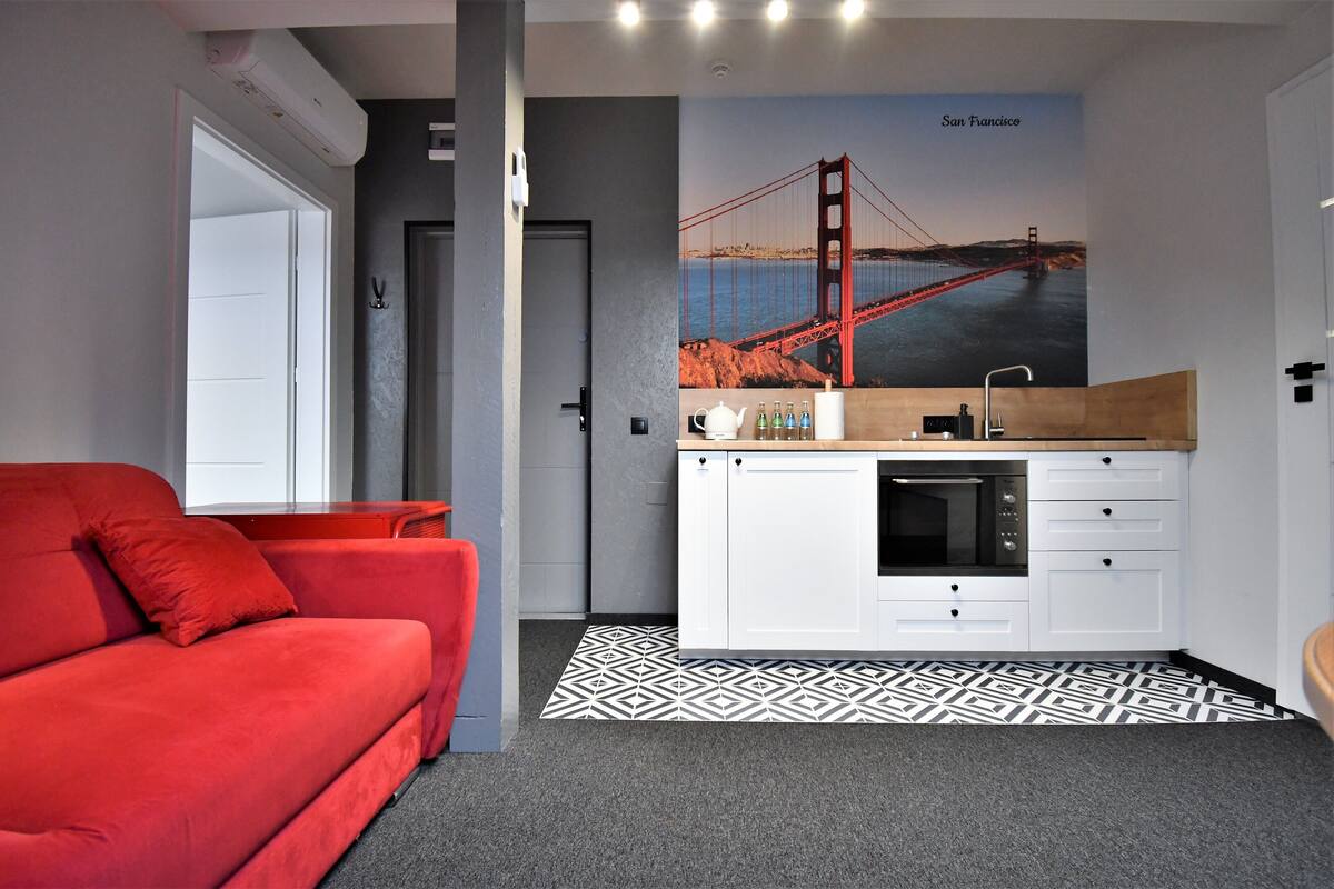 Una habitación moderna con un sofá rojo, una cocina con armarios blancos y un mural grande del Puente Golden Gate.