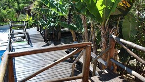 A wooden deck with a metal railing, surrounded by banana trees and a pool. - Pousada Bezerra (Nazare Paulista)