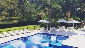 A rectangular swimming pool with a tiled edge, surrounded by white lounge chairs and umbrellas, set against a backdrop of greenery and trees. - Pousada Bezerra (Nazare Paulista)