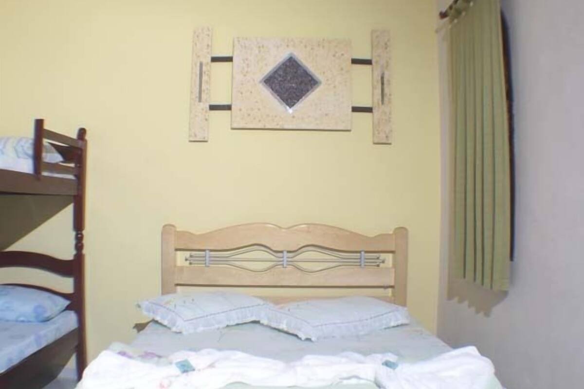 Un lit simple avec une tête de lit en bois, un élément décoratif fixé au mur au-dessus, et une fenêtre avec des rideaux.
