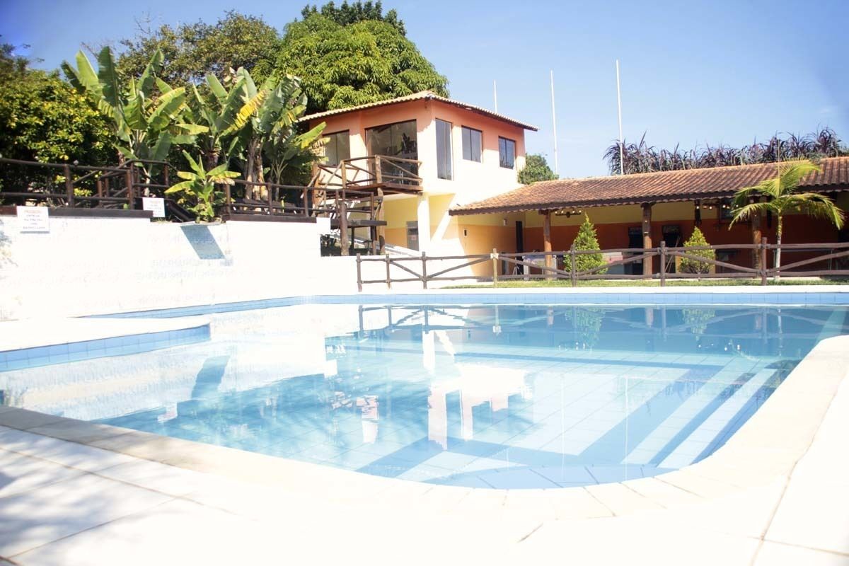 Uma piscina com deck de madeira, um edifício com telhado vermelho e vegetação exuberante ao fundo.