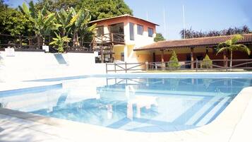Uma piscina com deck de madeira, um edifício com telhado vermelho e vegetação exuberante ao fundo.