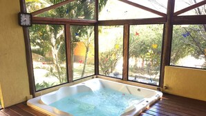 A hot tub with a wooden deck, surrounded by large windows with a view of greenery. - Pousada Bezerra (Nazare Paulista)