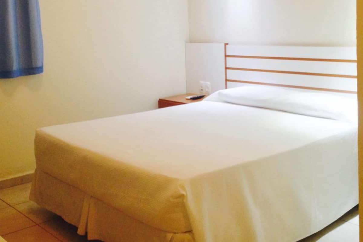 A simple, clean hotel room with a single bed, a nightstand, and a plain wall.