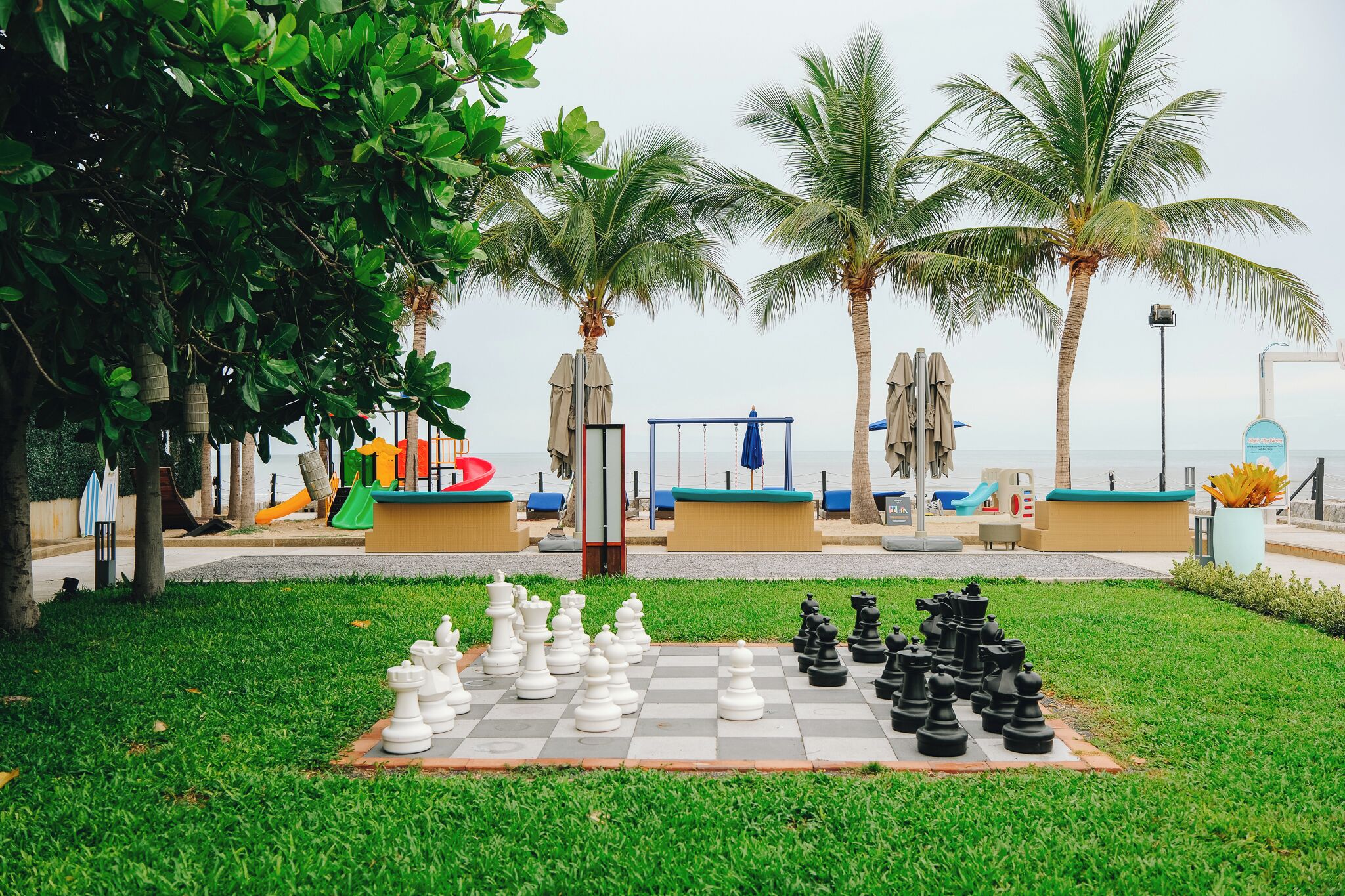 A chessboard set up outdoors with palm trees and beach chairs in the background.