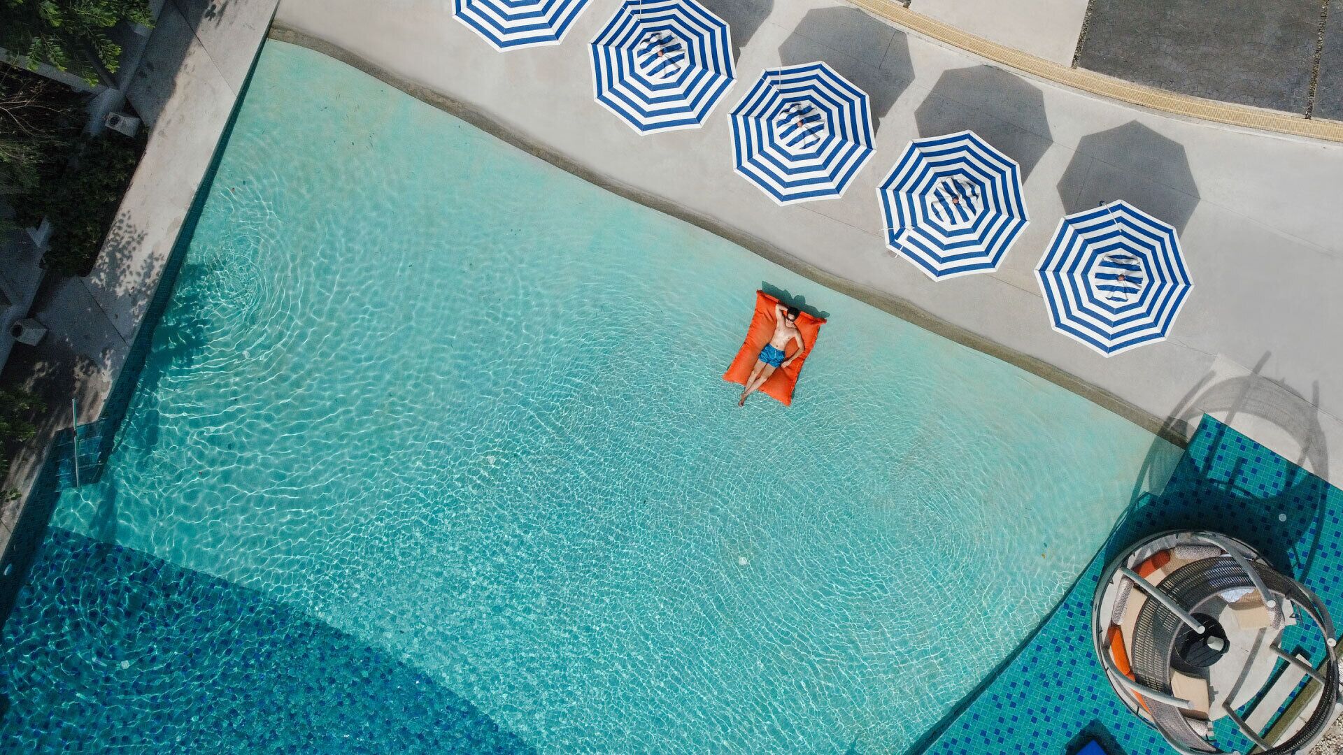 Une vue aérienne d’une piscine à l’eau claire, entourée de parasols à rayures bleues et blanches, et d’un espace aménagé au bord de la piscine avec une chaise et une structure circulaire.