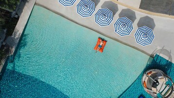 Vista aérea de una piscina con agua cristalina, rodeada de sombrillas azul y blanco, y un área de piscina con una silla y una estructura circular.