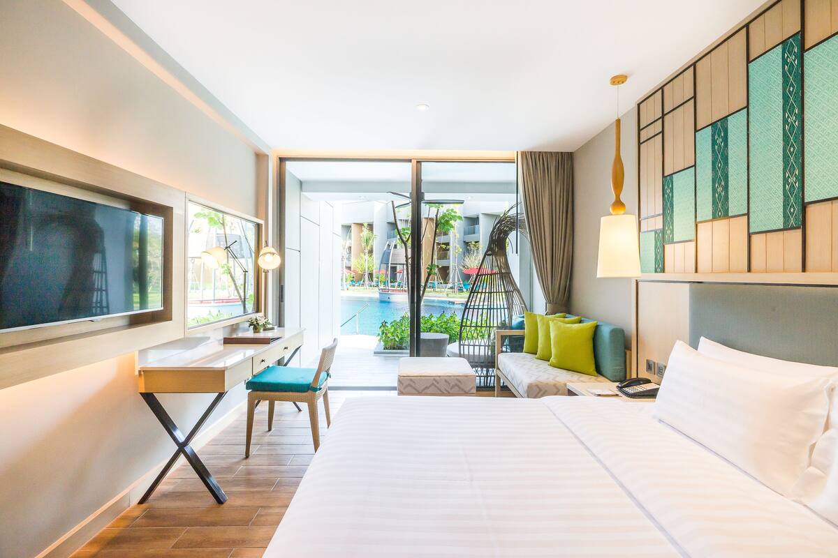 Een hotelkamer met een bed, bureau, stoel en uitzicht op een zwembad en gebouwen.