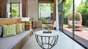 A modern living room with a sofa, a round coffee table, and a dining area with chairs. There is a large window providing a view of the garden. - Avani+ Hua Hin Resort (Cha-am)