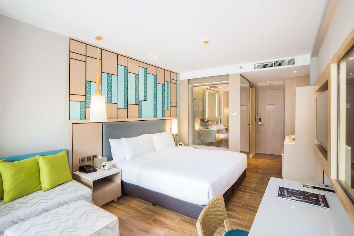 Een hotelkamer met een groot bed, een bureau en een zithoek. Er hangt een spiegel aan de muur en een deur naar de badkamer.