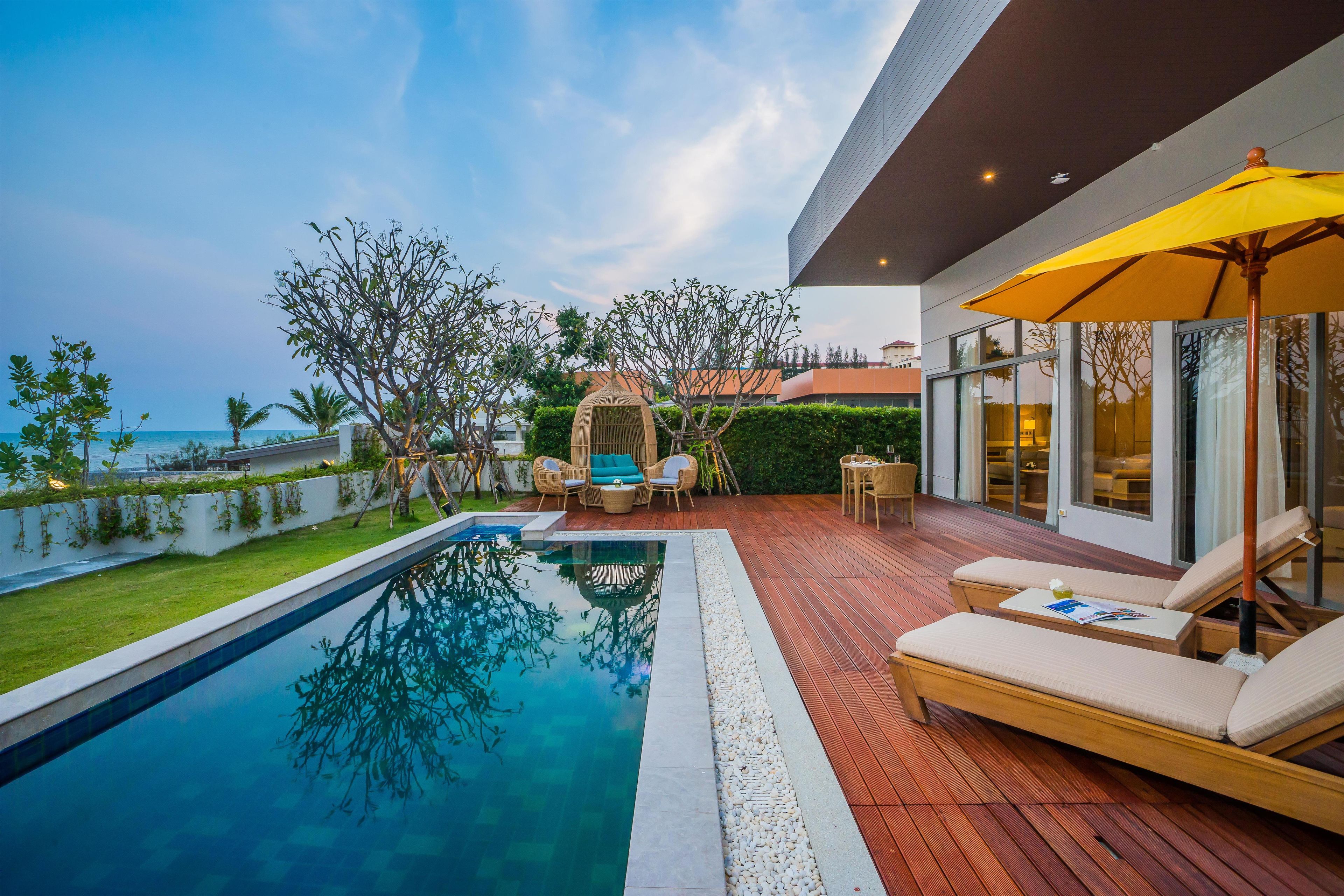 A modern outdoor pool area with a wooden deck, lounge chairs, and a view of the sea.