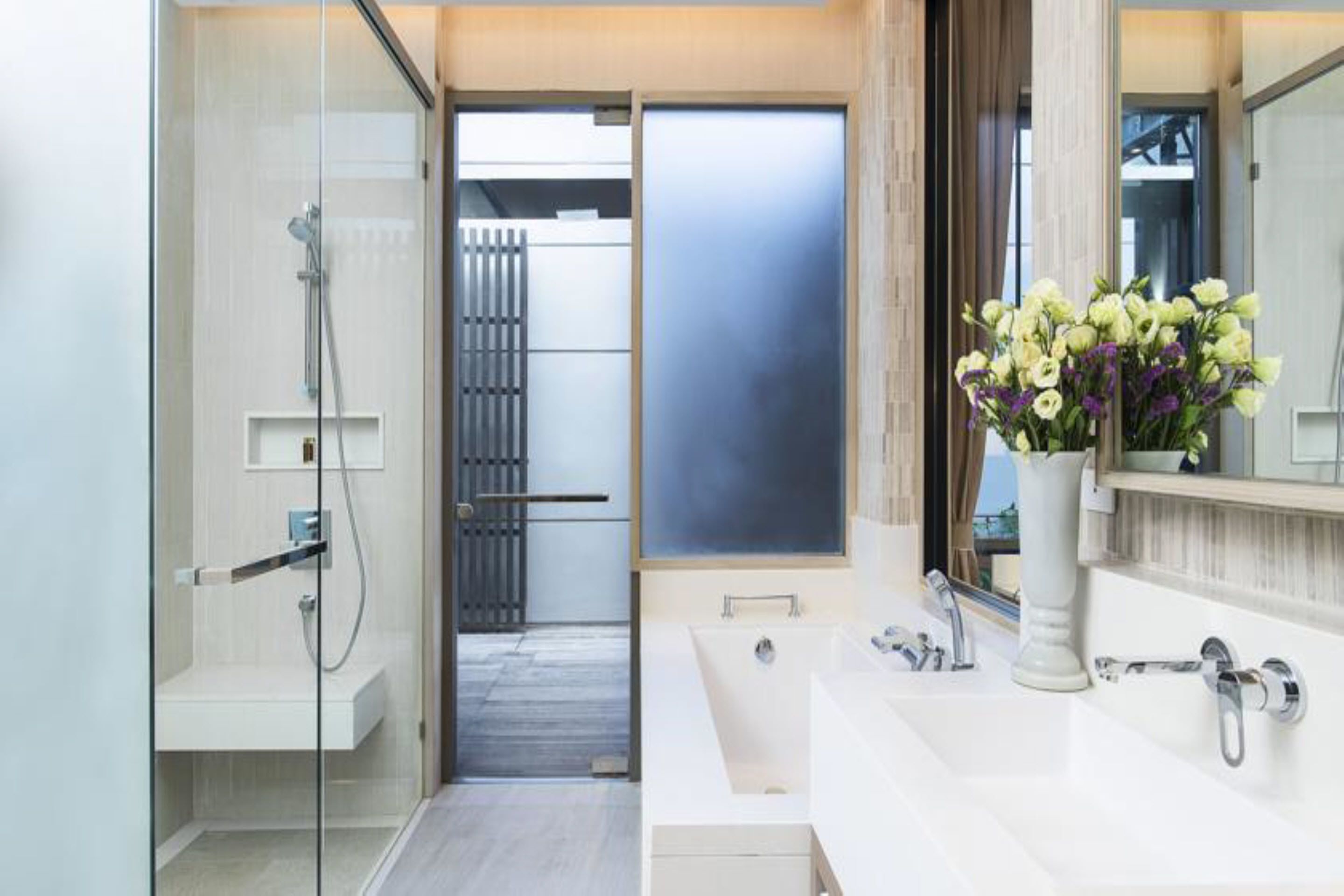 A modern bathroom with a glass shower enclosure, a large mirror, and a vase of flowers on the vanity.