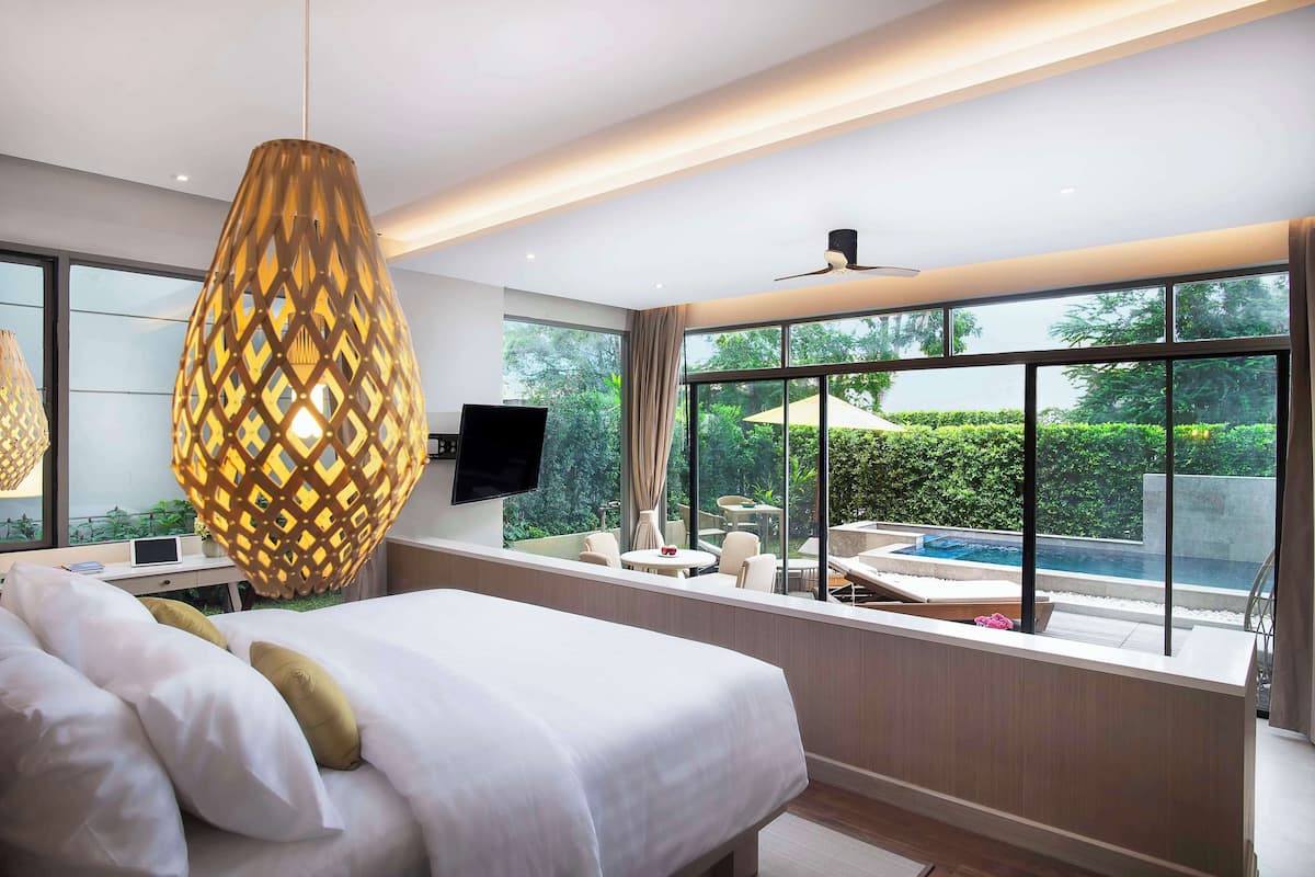 Een moderne hotelkamer met een groot bed, een bureau met een computer, een televisie en uitzicht op een zwembad en groen.