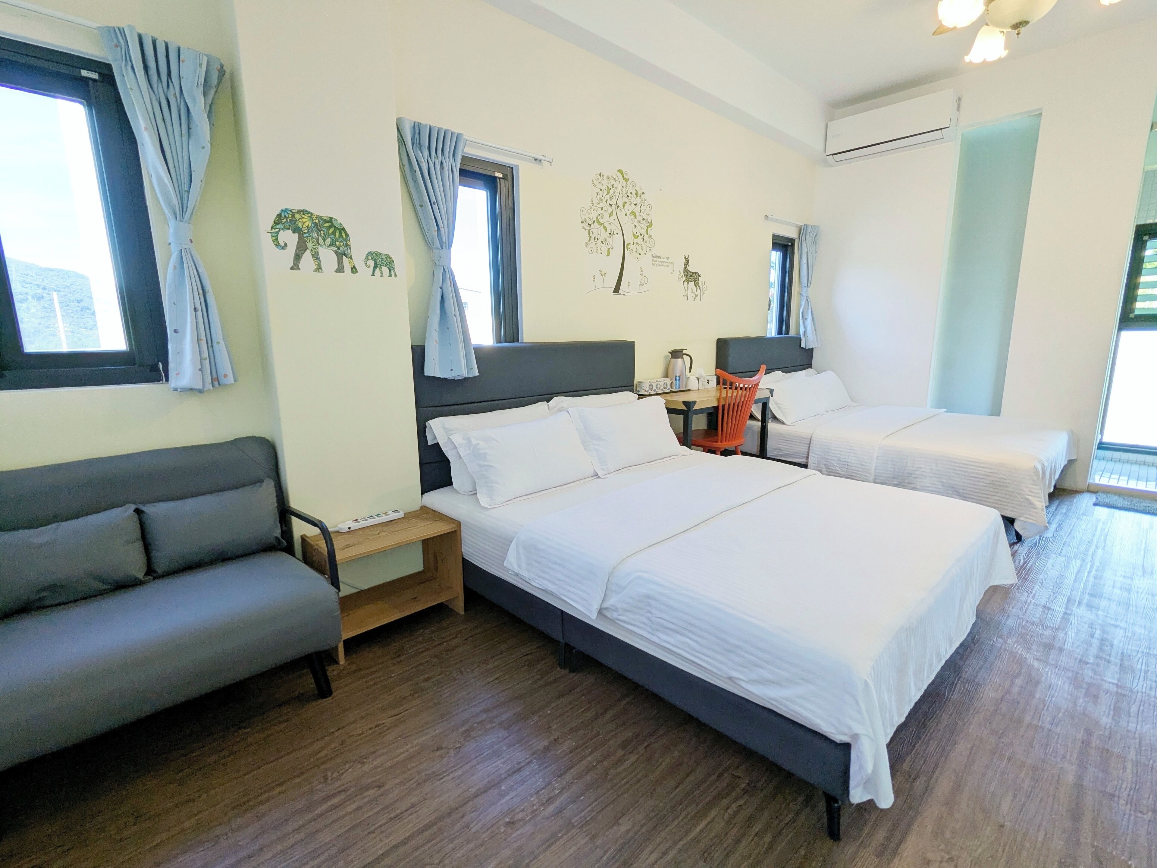 A hotel room with two beds, a sofa, a table with a kettle, and a chair.