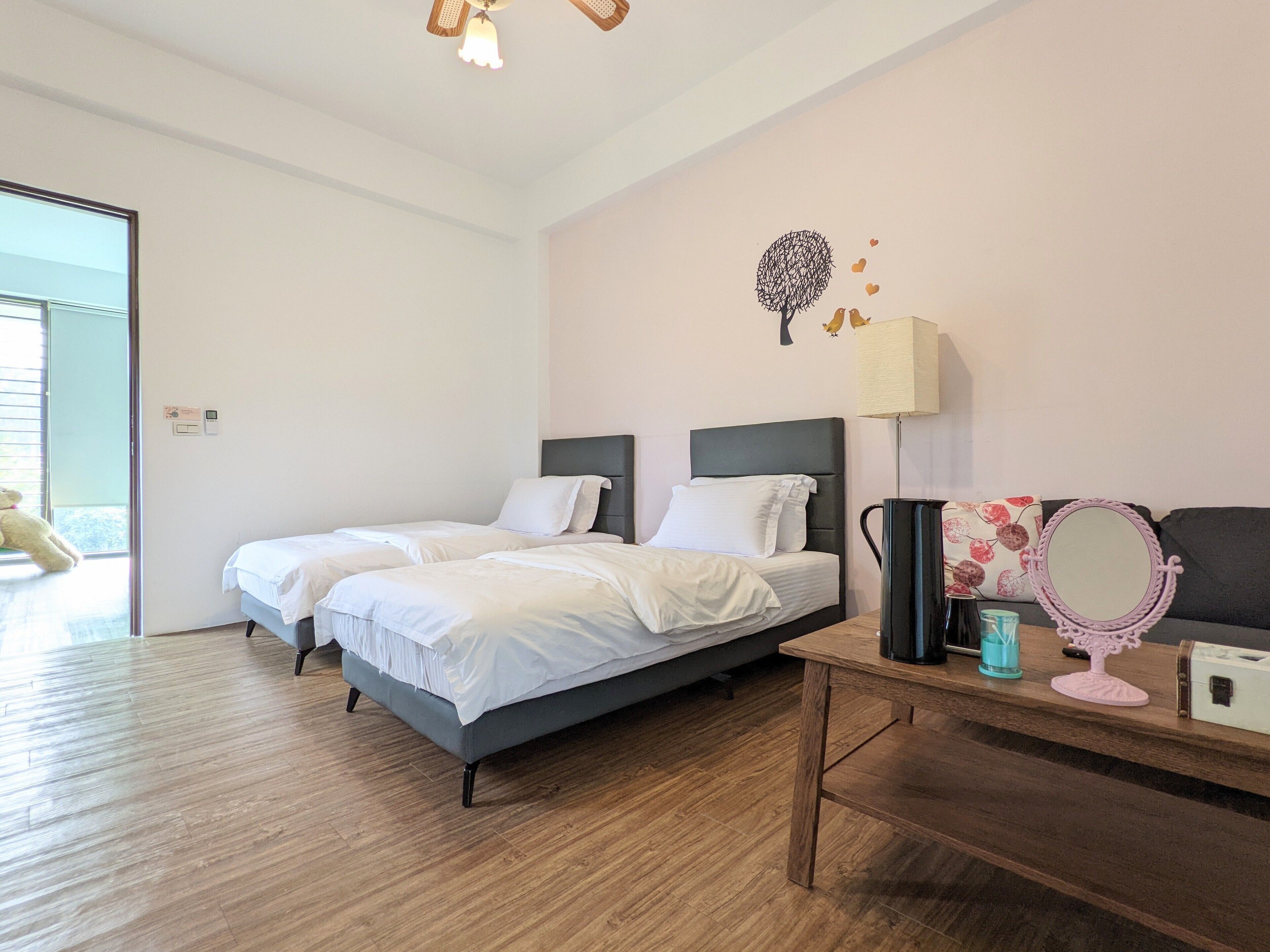 A bedroom with two beds, a wooden floor, a bedside table with a lamp, a mirror, and a wall decoration.