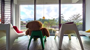 Una habitación con un ventanal grande, un oso de peluche sobre un taburete verde y sillas blancas.