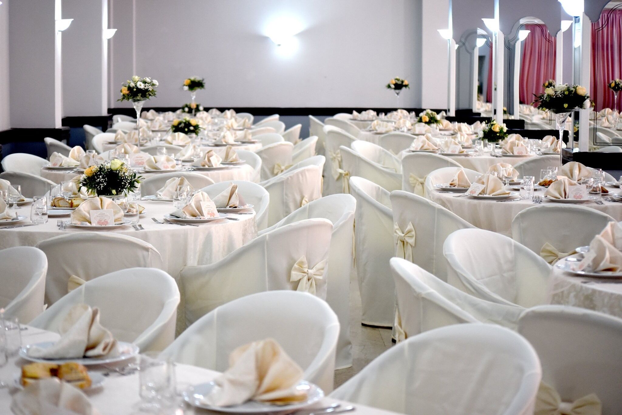 Ein Festsaal mit runden Tischen, gedeckten mit weißen Tischdecken, Stühlen mit weißen Bezügen und floralen Tischdekorationen.