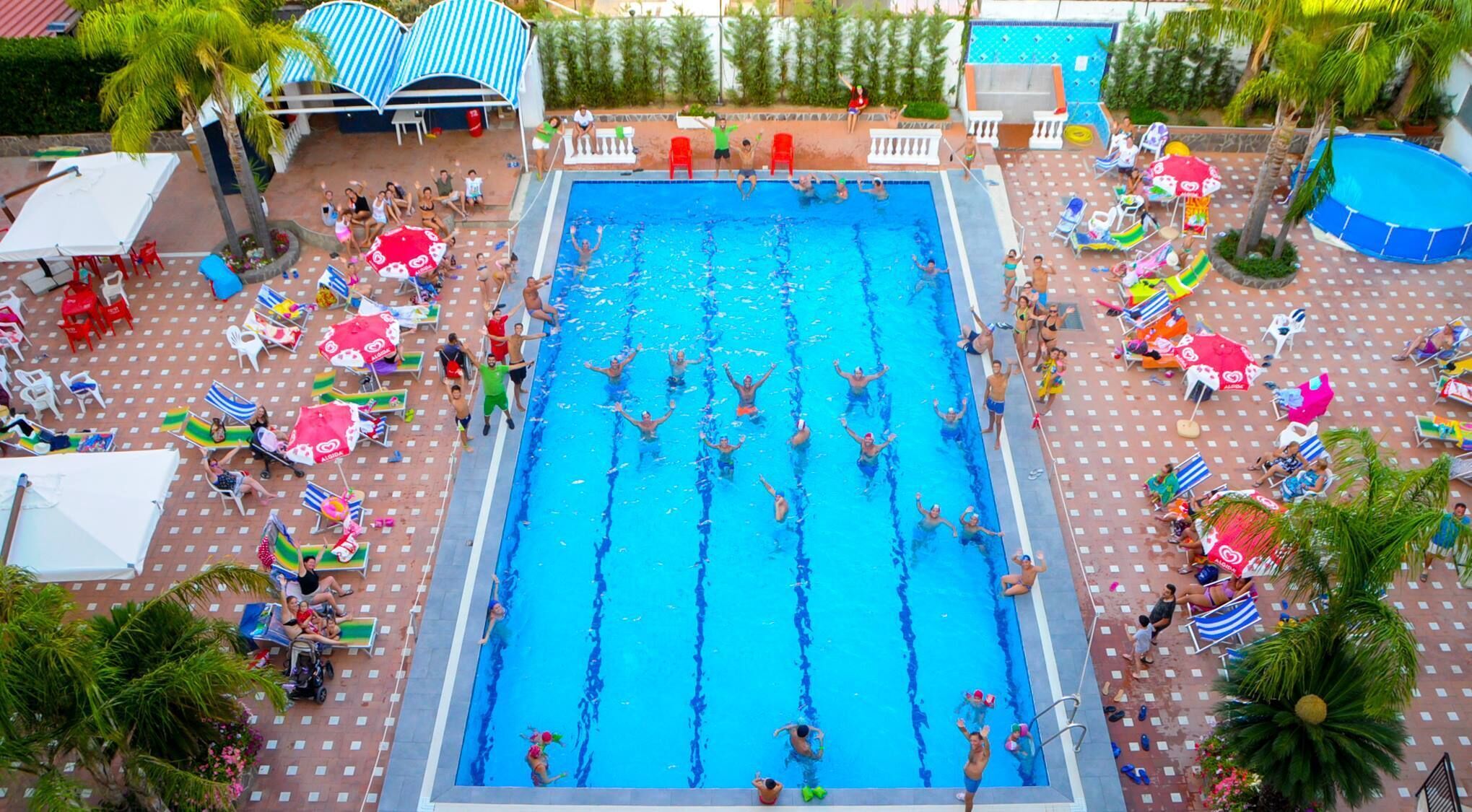 A swimming pool with multiple lanes, surrounded by sun loungers and umbrellas, with palm trees and a covered seating area in the background.