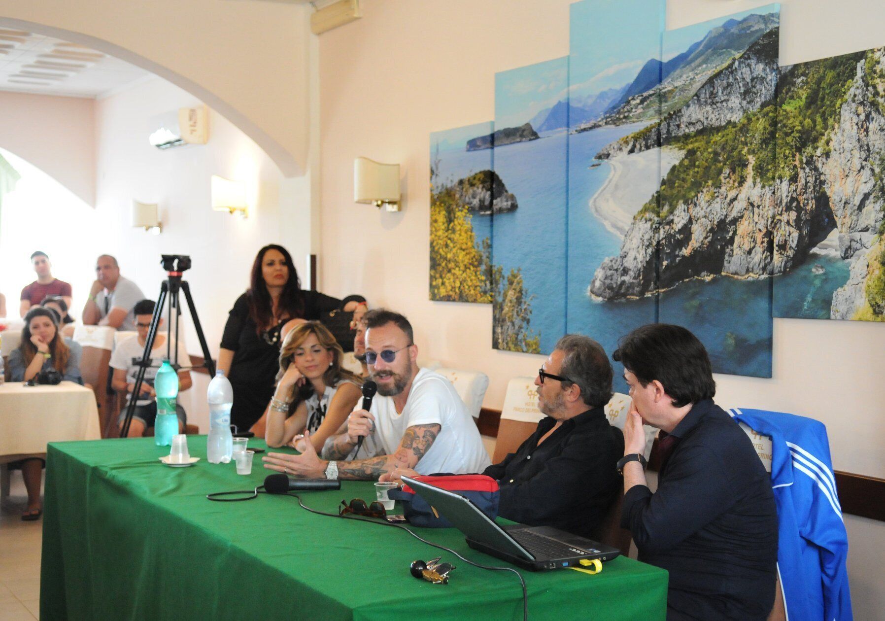 Uma sala de conferências com mesa verde, laptops e microfones, e uma parede com uma pintura da paisagem de montanhas.