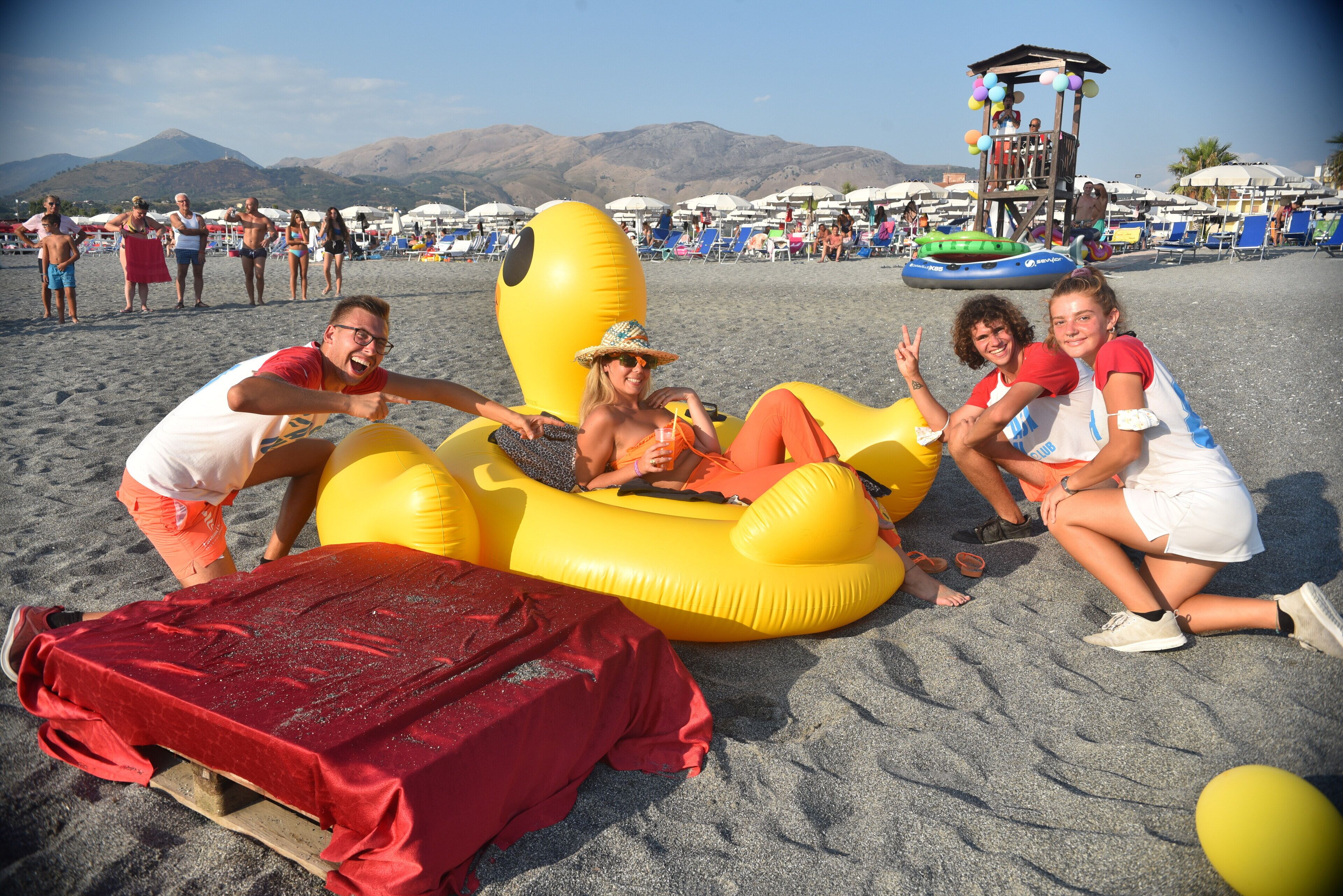 Escena de playa con balsas inflables, una torre de salvavidas y gente en la playa.