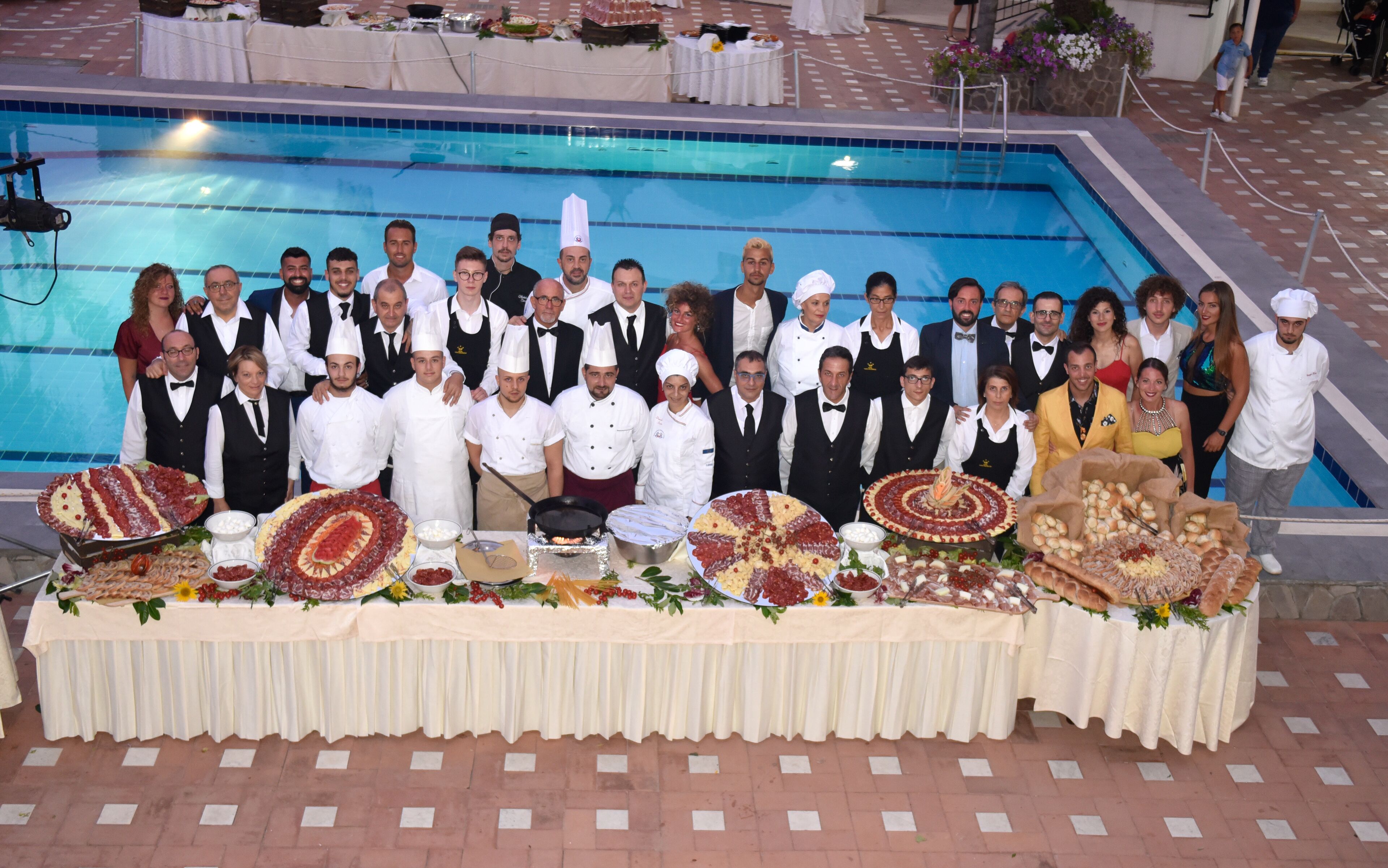 A group of people posing by a poolside buffet with various food items.