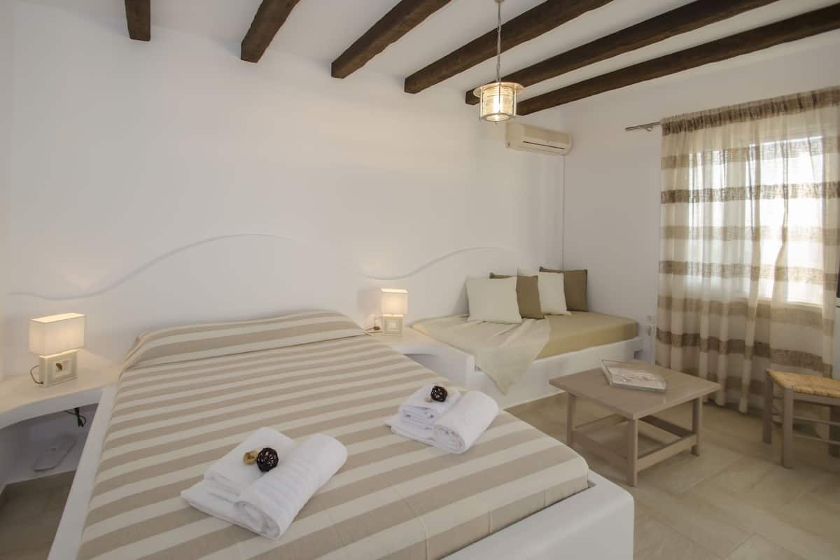 A hotel room with a large bed, a smaller bed, a bedside table with a lamp, a bench, and a window with curtains.