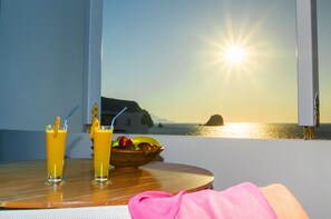 Una mesa con dos vasos de jugo de naranja y un cuenco de fruta, con vista a una puesta de sol costera.
