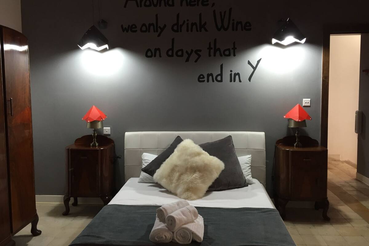 Un dormitorio con cama, dos mesitas de noche con lámparas, una pared con una cita motivacional y una puerta a la derecha.