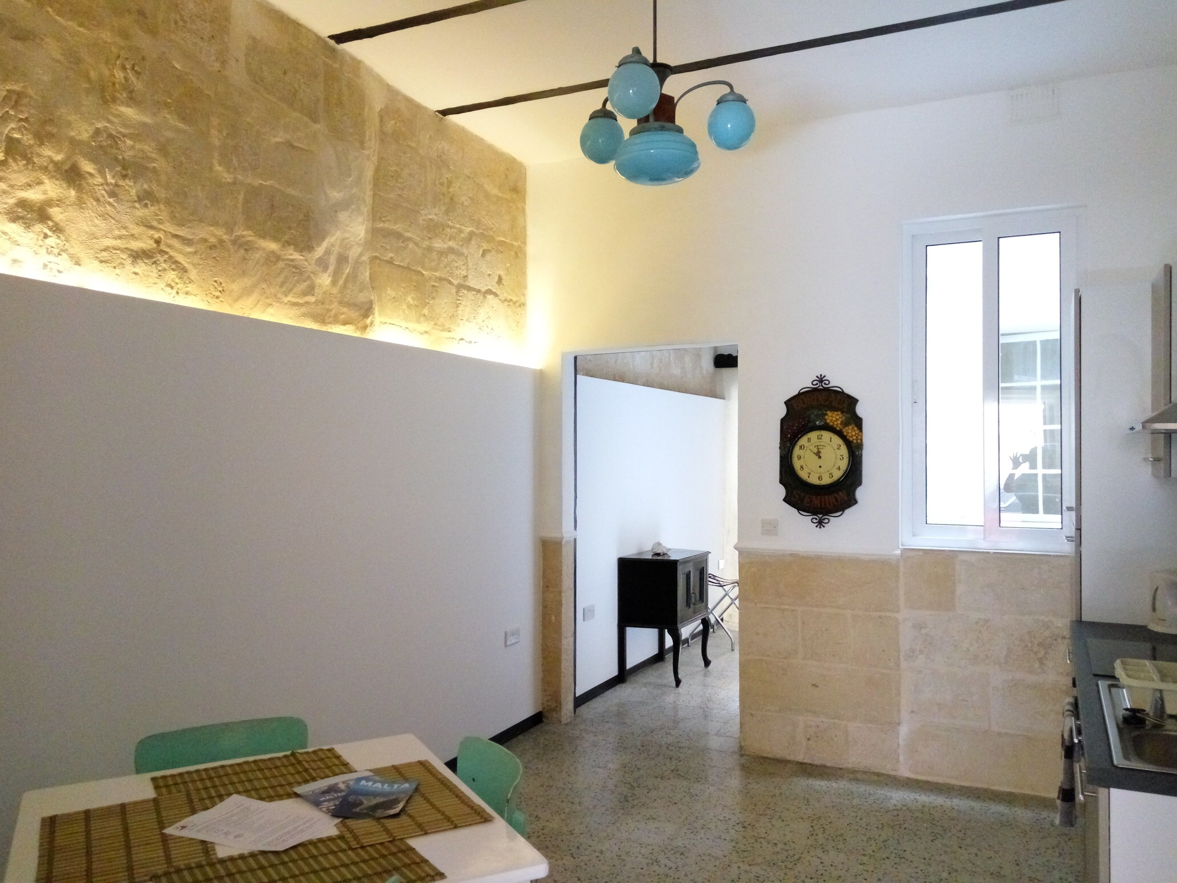 A dining area with a table set for two, a clock on the wall, and a kitchen area visible through an open door.
