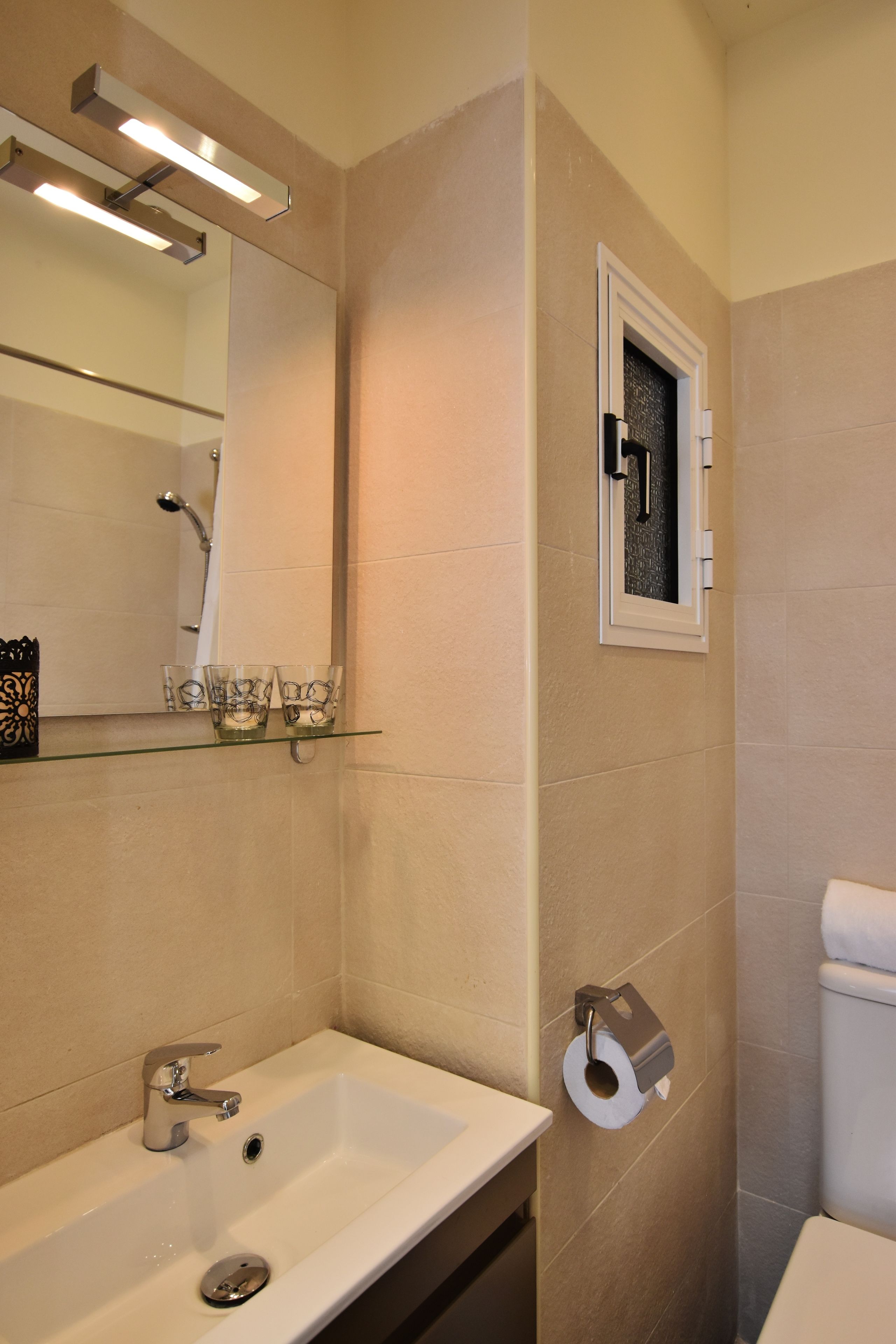 A bathroom with a white sink, a mirror, a toilet, and a glass shelf with cups.