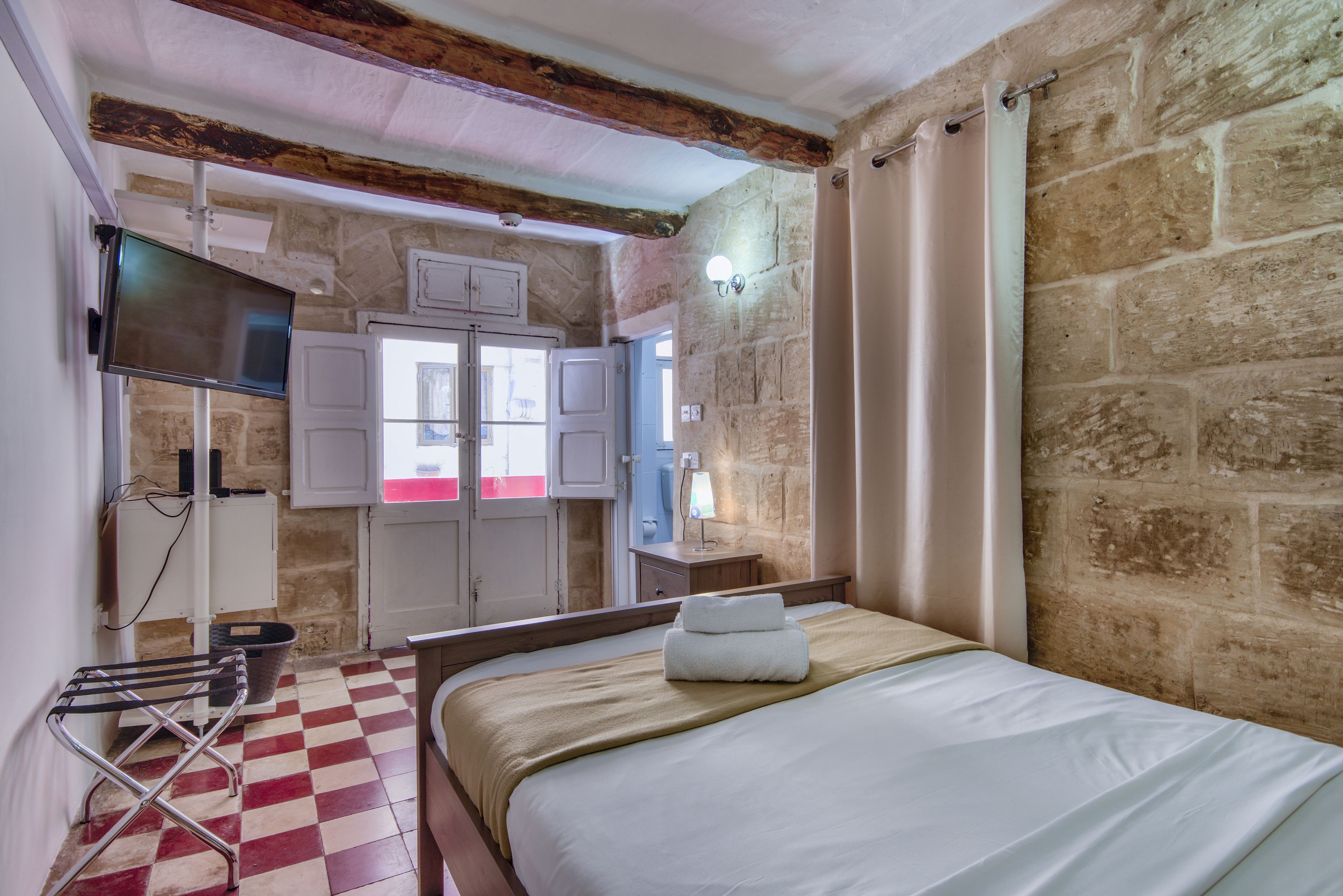 A hotel room with a checkered floor, a bed with white linens, a flat-screen TV mounted on the wall, a wooden ceiling, stone walls, and a window with curtains.