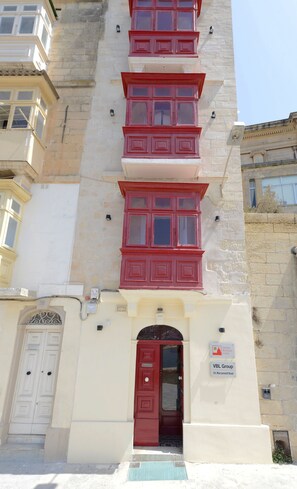 Un bâtiment avec une porte rouge et de nombreuses fenêtres. On y voit une enseigne indiquant « Valette Boutique Living » et « VBL Group », ainsi qu’une adresse.