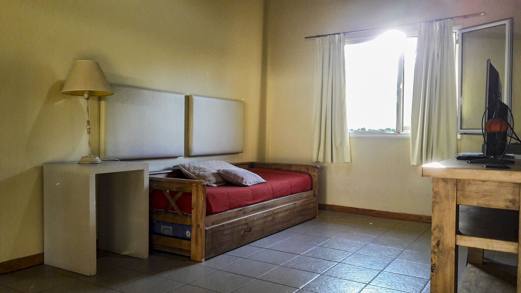 Ein kleines Zimmer mit Bett, Tisch, Lampe und einem Fenster mit Vorhängen.