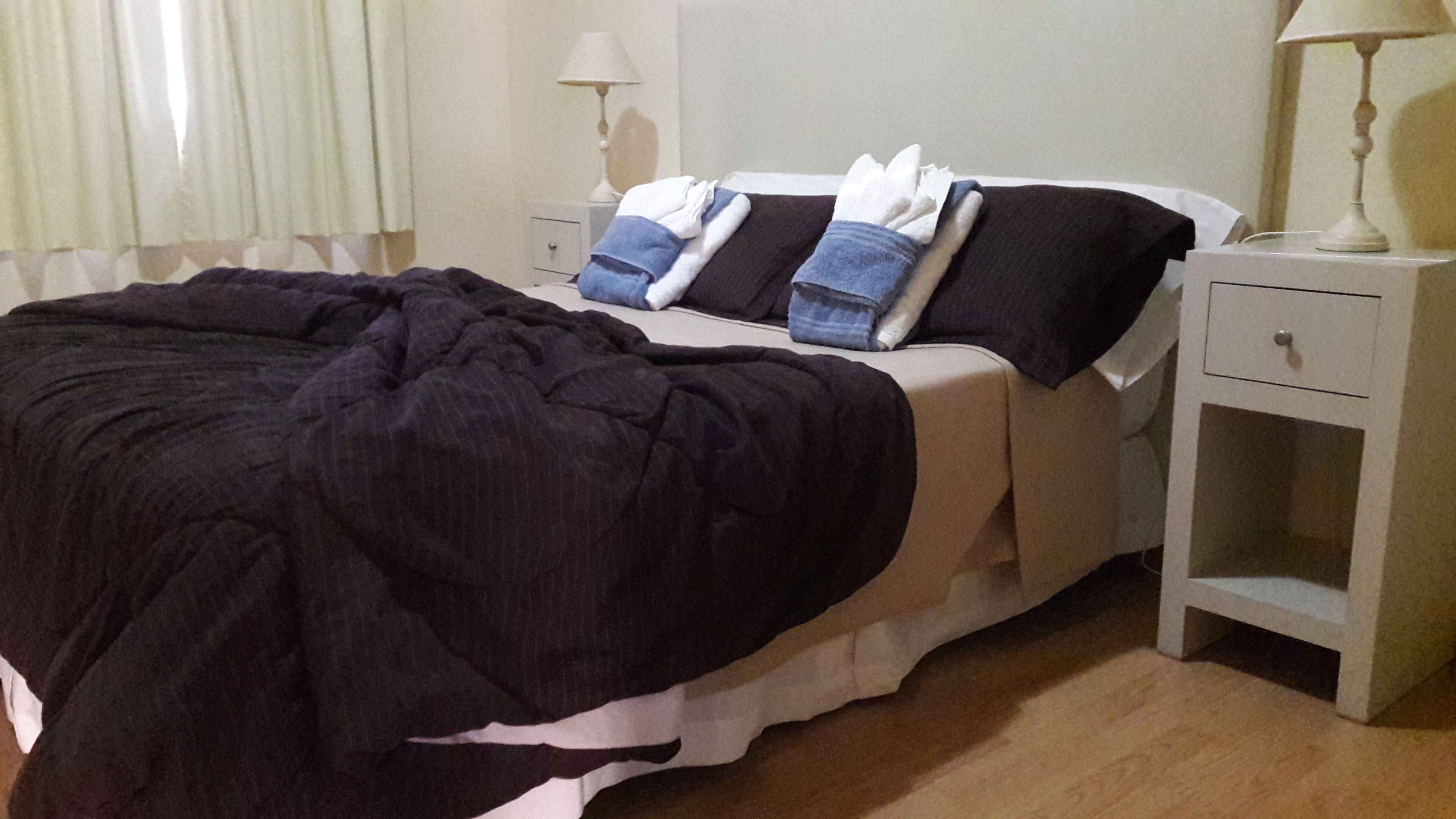 Cama bem arrumada com edredom escuro e almofadas, mesa de cabeceira com luminária e um móvel com gavetas.