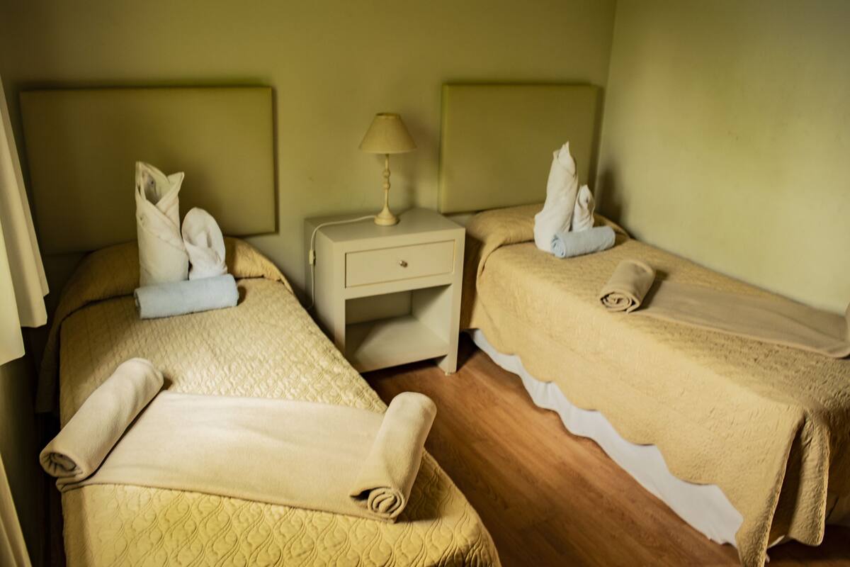 A hotel room with two beds, each with a pillow and a rolled towel. There is a nightstand with a lamp between the beds.