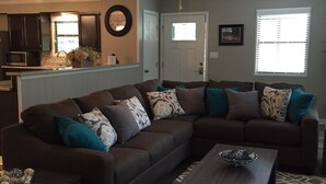 A living room with a sectional sofa, a coffee table, a patterned rug, a ceiling fan, and a kitchen area in the background. - Whitewater Condos (Canyon Lake)